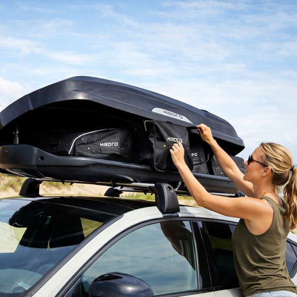 Hapro dakkoffertassen laden op auto – ideaal voor vakantie en roadtrip Vrouw plaatst bagage in Hapro dakkoffer op auto bij strand – extra bagageruimte voor vakantie